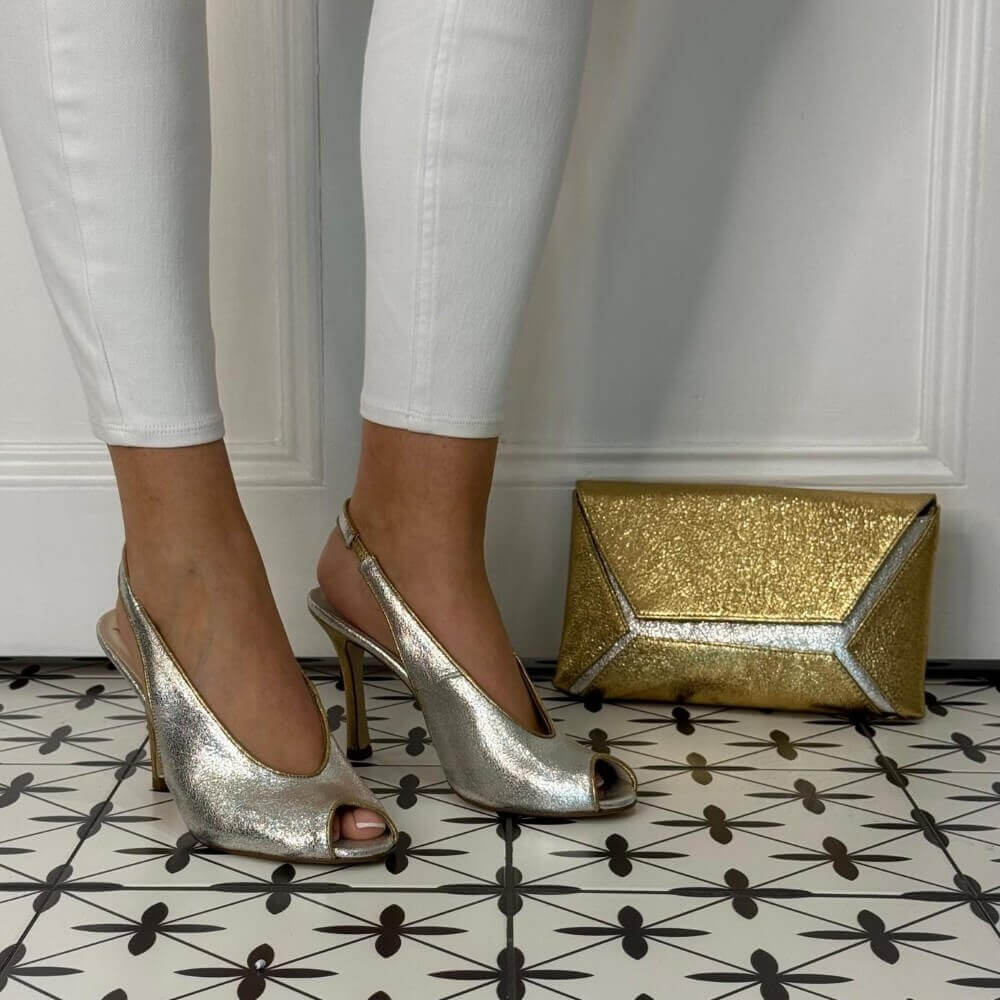 Silver high-heeled shoes worn with white pants on a patterned floor, next to a gold clutch.