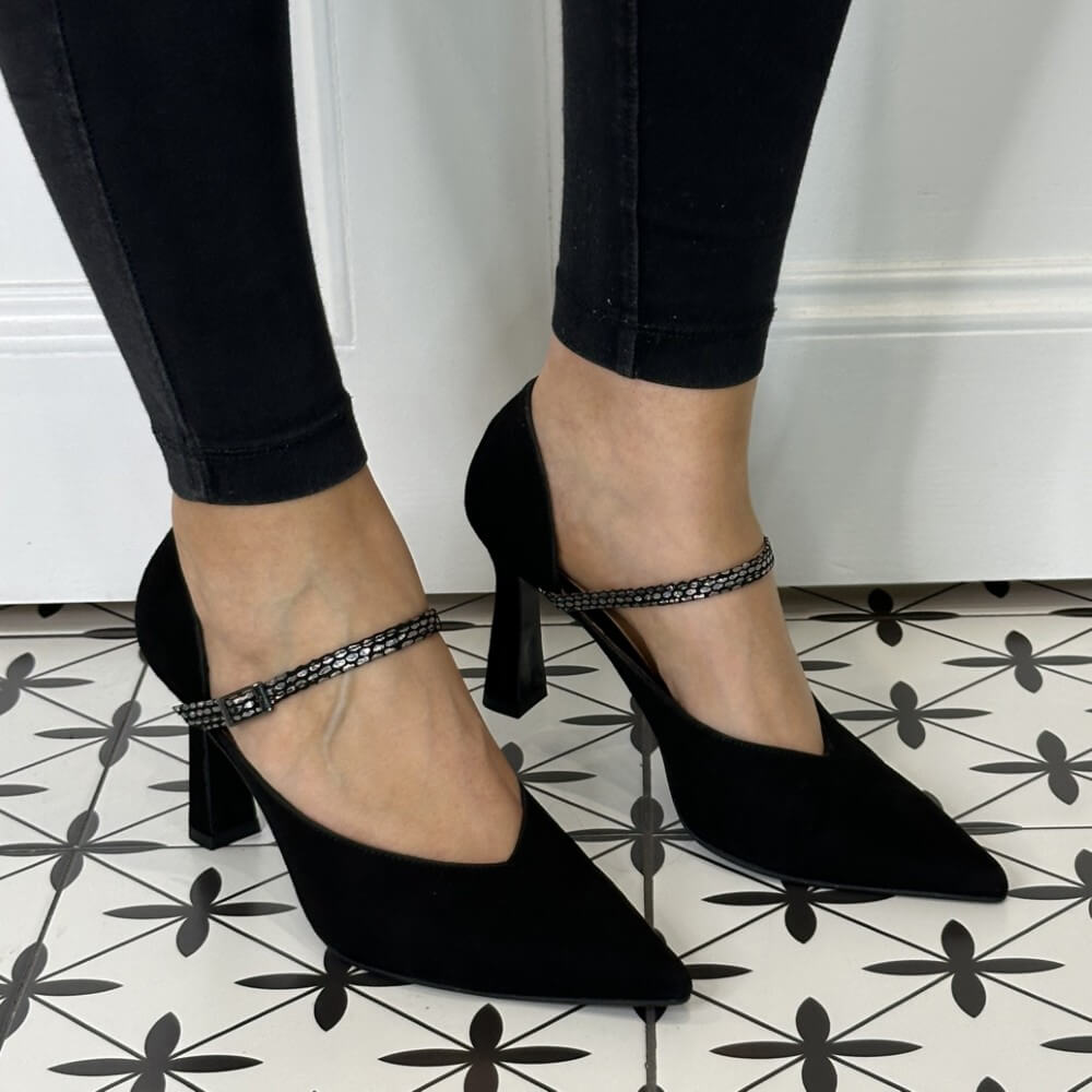 Black suede leather high-heeled shoes with ankle straps worn by a person on a patterned floor.