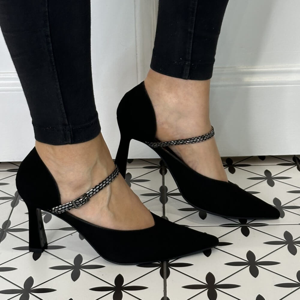 Black suede leather high-heeled shoes with ankle straps worn by a person on a patterned floor.