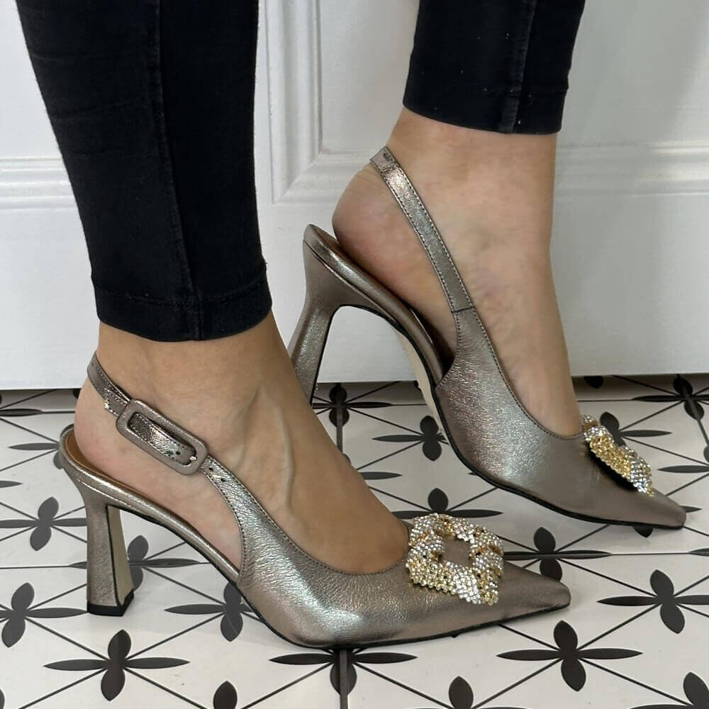 Gold high-heeled shoes with embellishments worn by a person on a patterned floor.