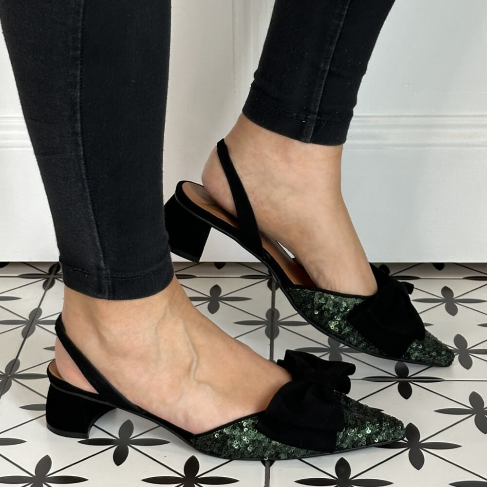 Black block-heeled shoes with glittery bow detail worn by a person on a patterned floor.