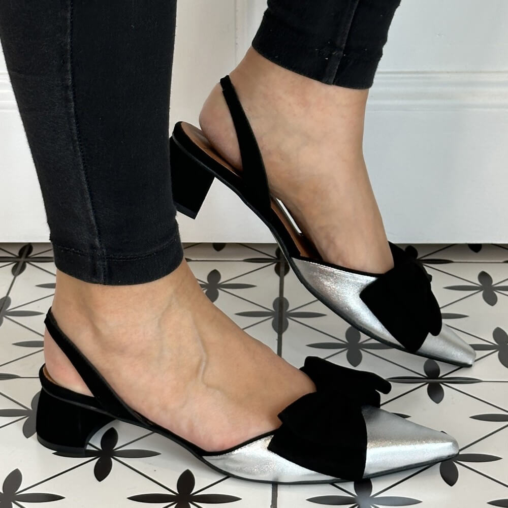 Silver block-heeled shoes with black bows worn by a person on a patterned floor.