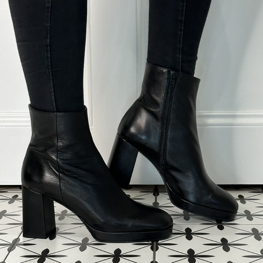 Black leather ankle boots worn with black pants on a patterned floor.