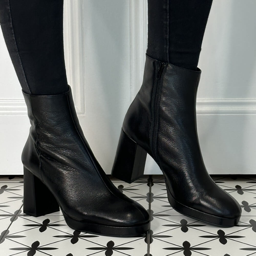 Black leather ankle boots worn with black pants on a patterned floor.