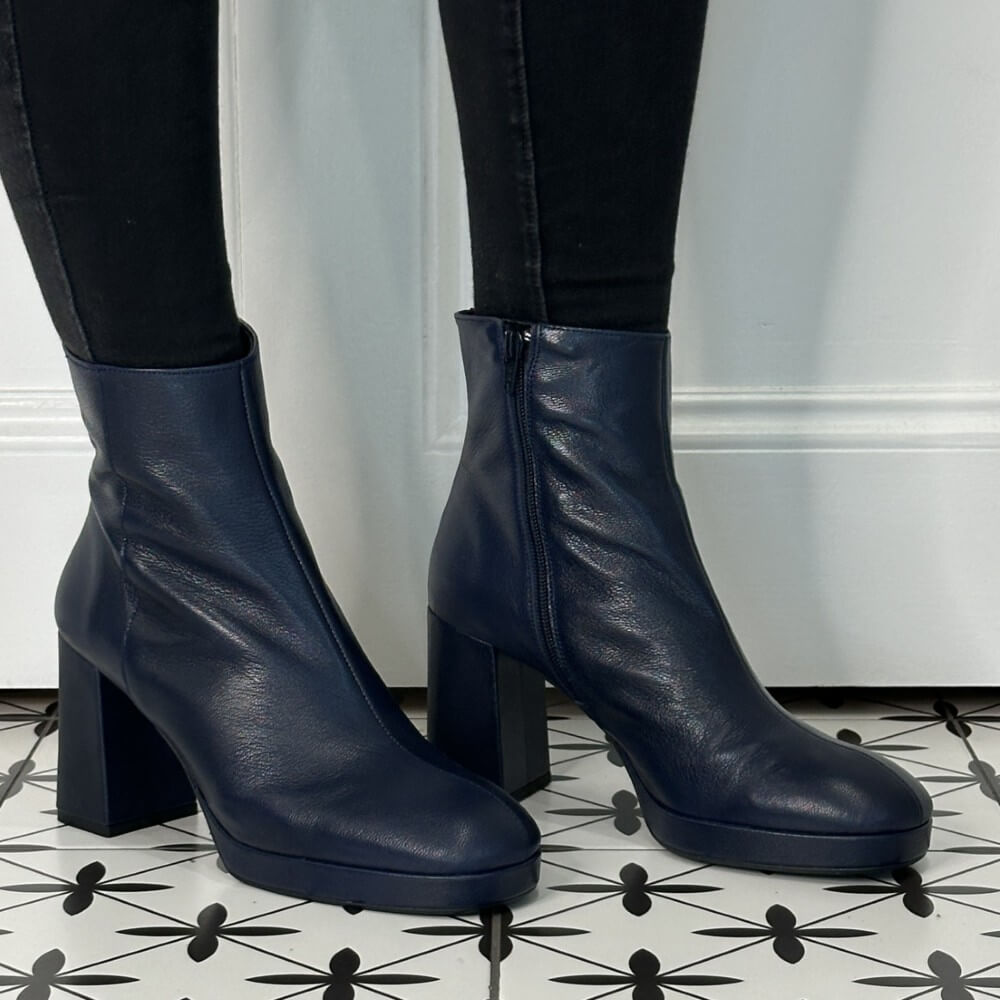 Navy blue leather ankle boots worn with black pants on a patterned floor.