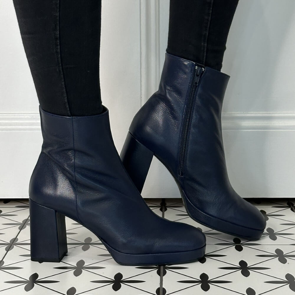 Navy blue leather ankle boots worn with black pants on a patterned floor.
