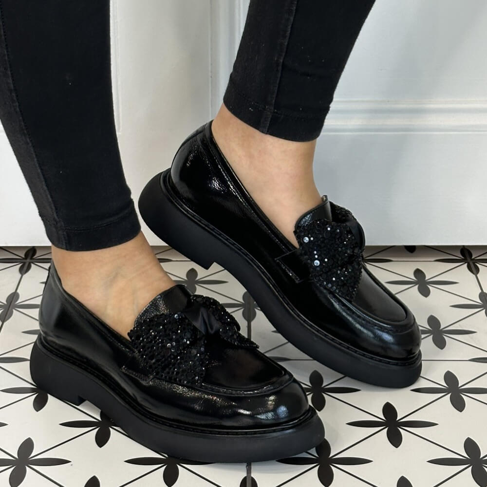 Black loafers with Diamante bow embellishments worn by a person on a patterned floor.