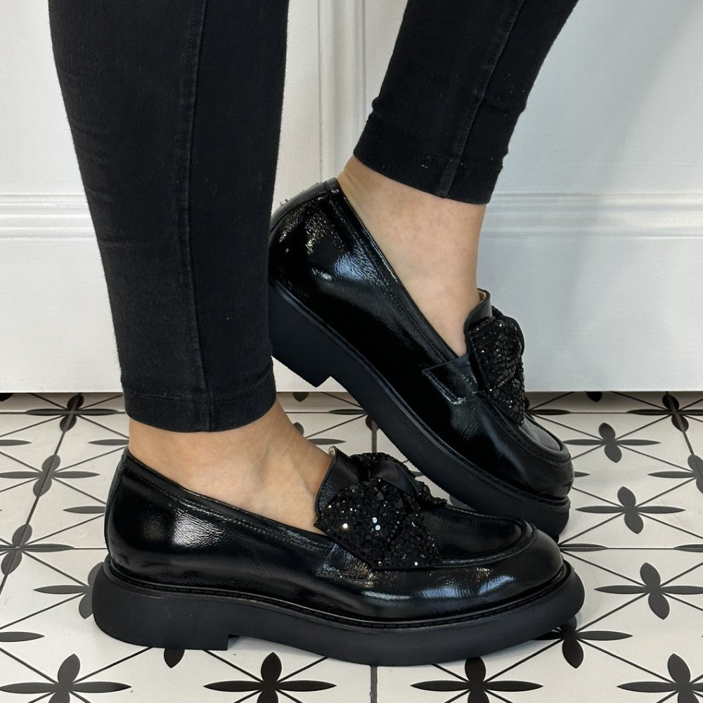 Black loafers with decorative bows worn by a person on a patterned floor.