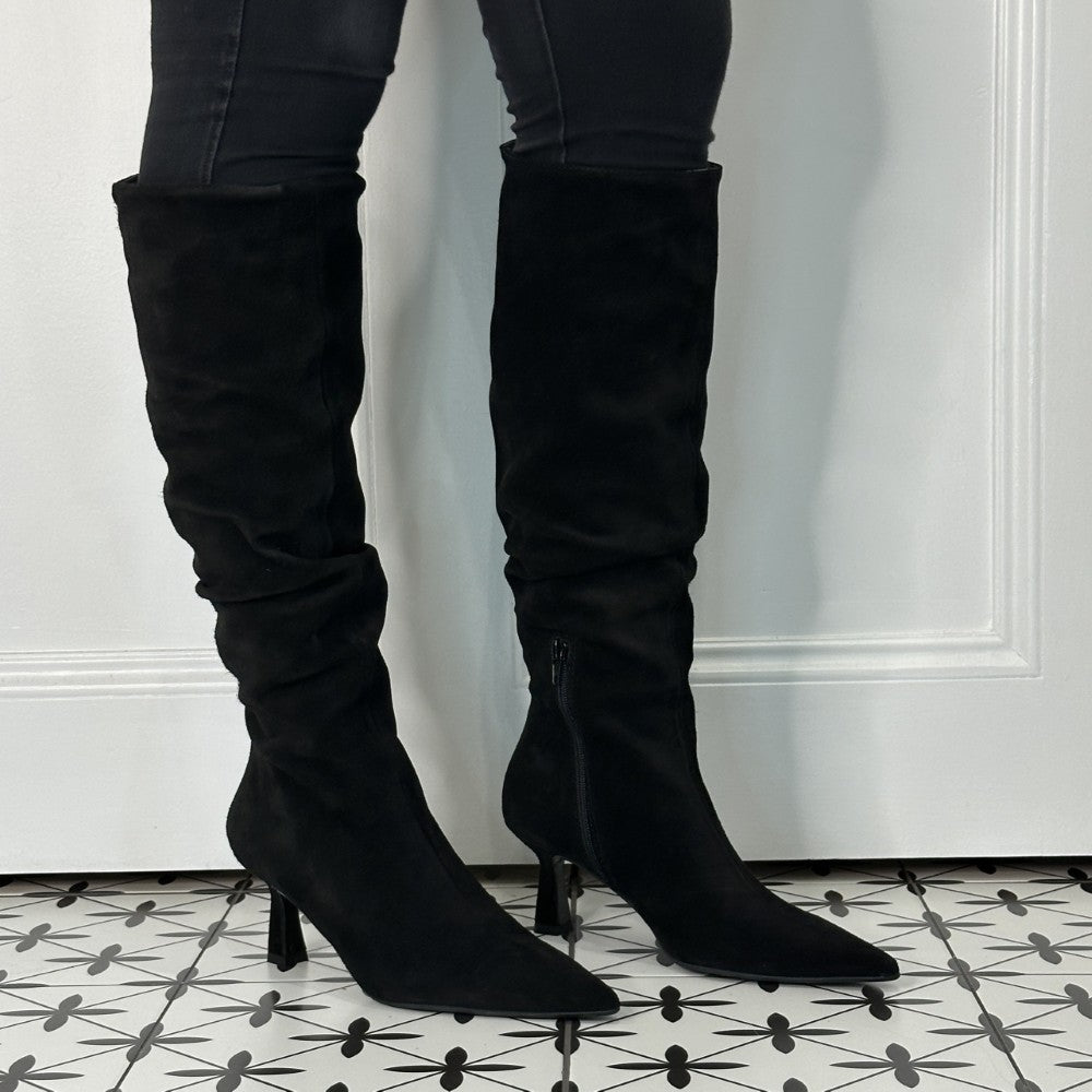 Black suede knee-high boots worn with black pants on a patterned floor.