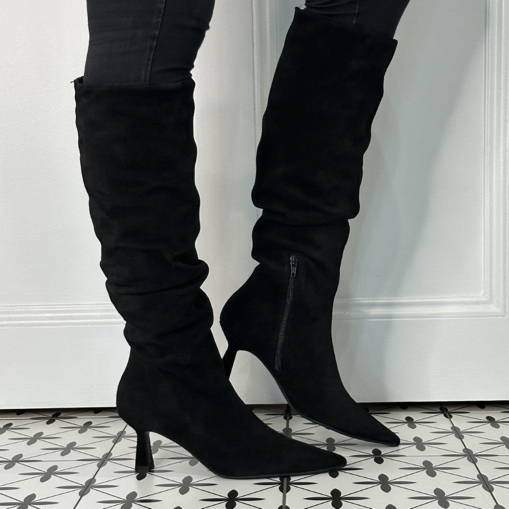 Black suede knee-high boots worn with black pants on a patterned floor.