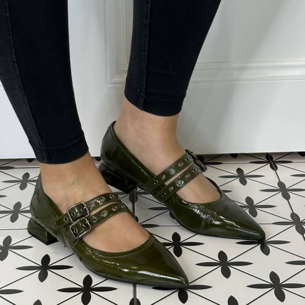 Green patent leather shoes with buckles worn by a person on a black and white patterned floor.
