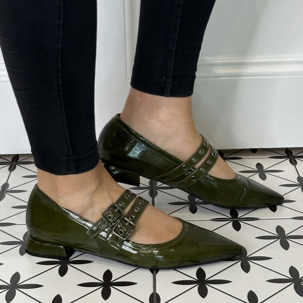 Green patent leather shoes with buckles worn by a person on a black and white patterned floor.