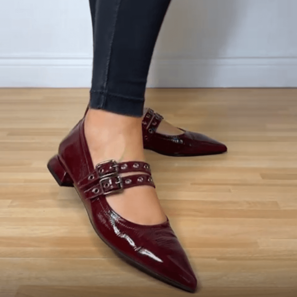 Burgundy pointed-toe shoes with buckles worn by a person on a wooden floor.