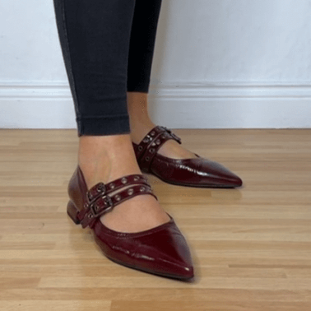 Burgundy pointed-toe shoes with buckles worn by a person on a wooden floor.