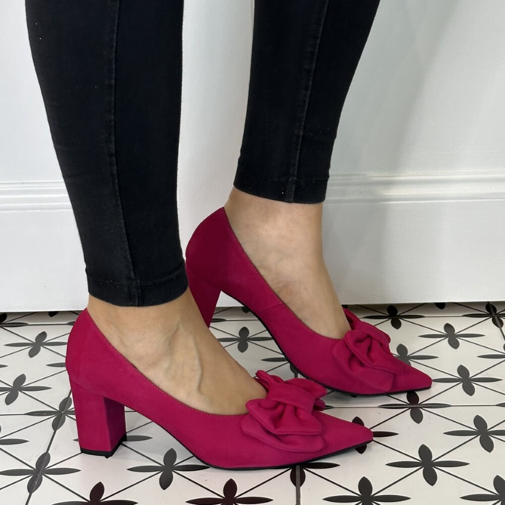 Pink high-heeled shoes with bows worn by a person on a patterned floor.