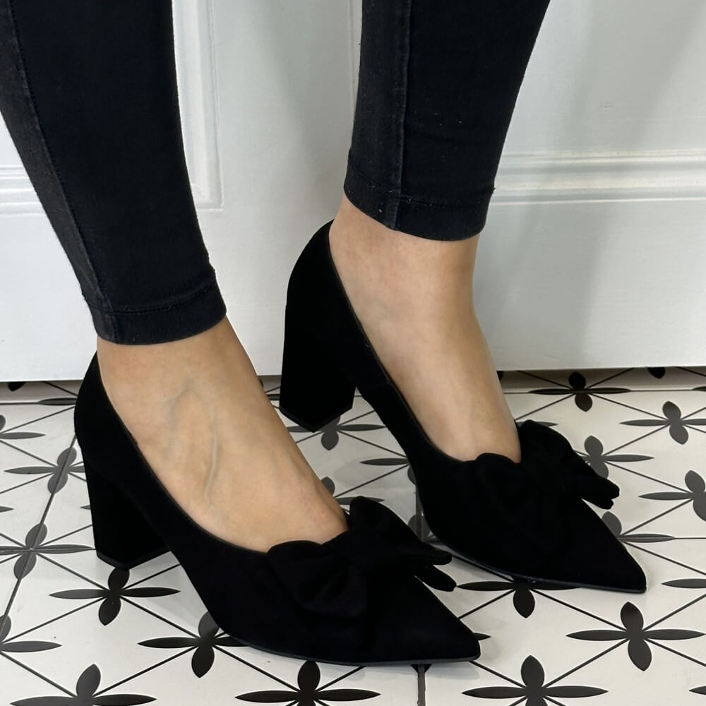 Black suede high heel shoes with bow details worn by a person on a patterned floor.
