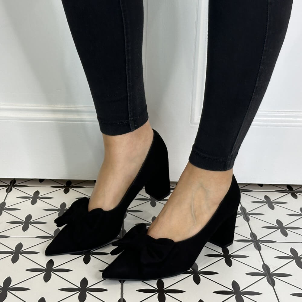 Black suede high heel shoes with bow details worn by a person on a patterned floor.