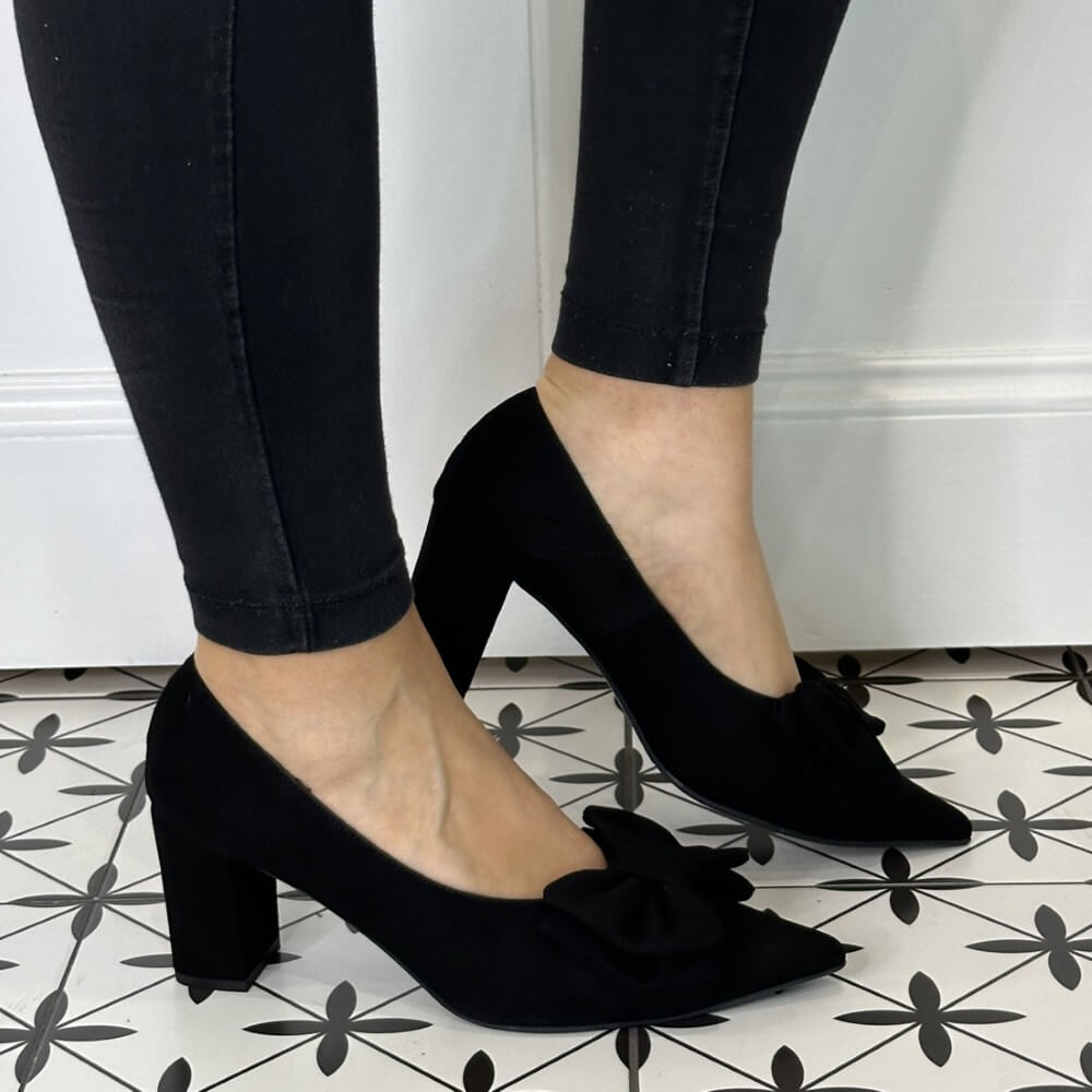 Black suede high-heeled shoes with a large bow, worn with black pants on a patterned floor.
