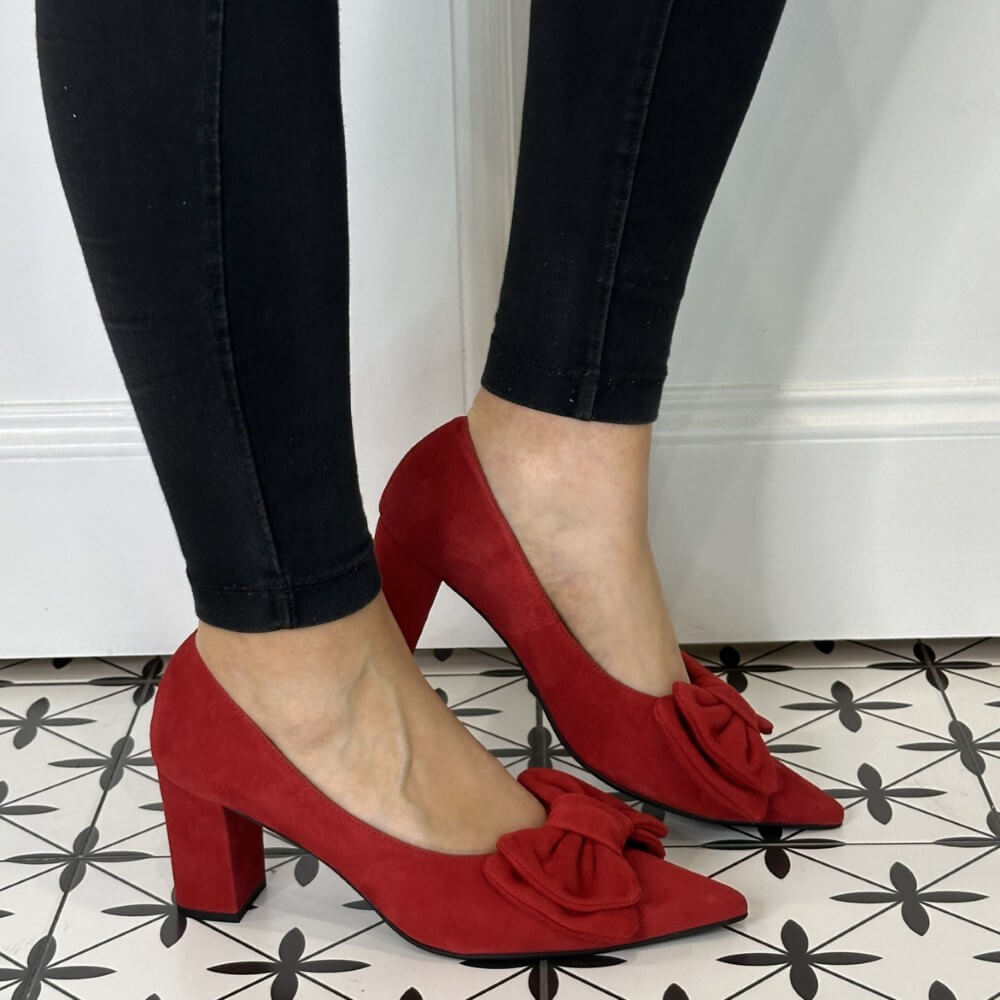 Red high-heeled shoes with bow details worn by a person on a patterned floor.