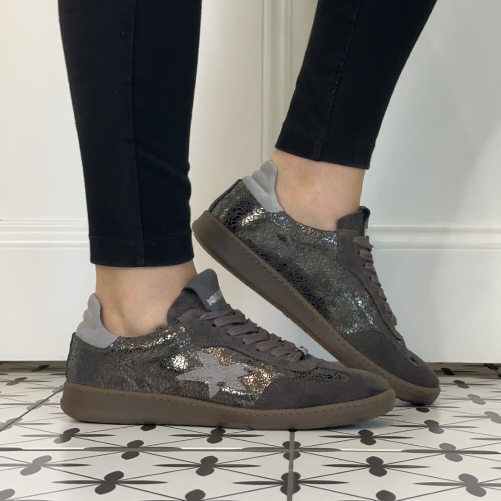 A pair of low-top Meline Retro sneakers with a dark grey glittery upper, brown suede-like overlays, and grey laces, shown on a person wearing black pants standing on a patterned tile floor.
