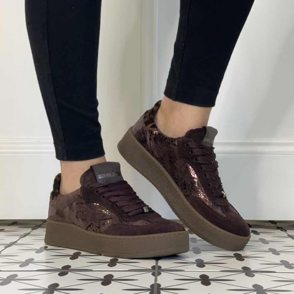A pair of Meline Retro low-top platform sneakers with dark brown suede and shimmering metallic sequins, featuring a leopard-print heel accent and thick brown soles, shown on a person wearing black pants.