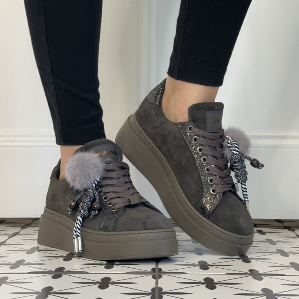 A person wearing Meline Retro dark grey suede platform sneakers with decorative tassels and a fluffy pom-pom, paired with black leggings, standing on a patterned tile floor.