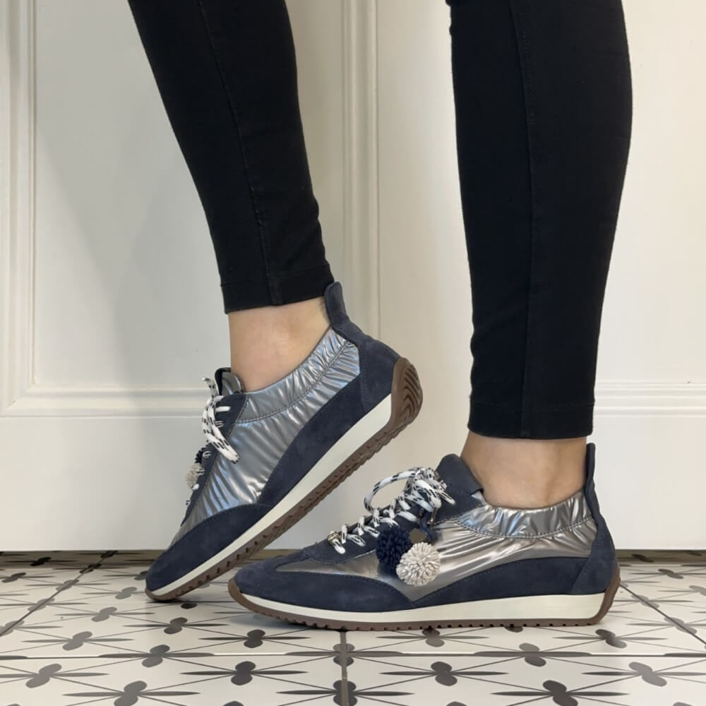 A person wearing Meline Retro navy and metallic silver low-top sneakers with white patterned laces, white pom-pom accents, and black leggings, standing on a black-and-white patterned tile floor.