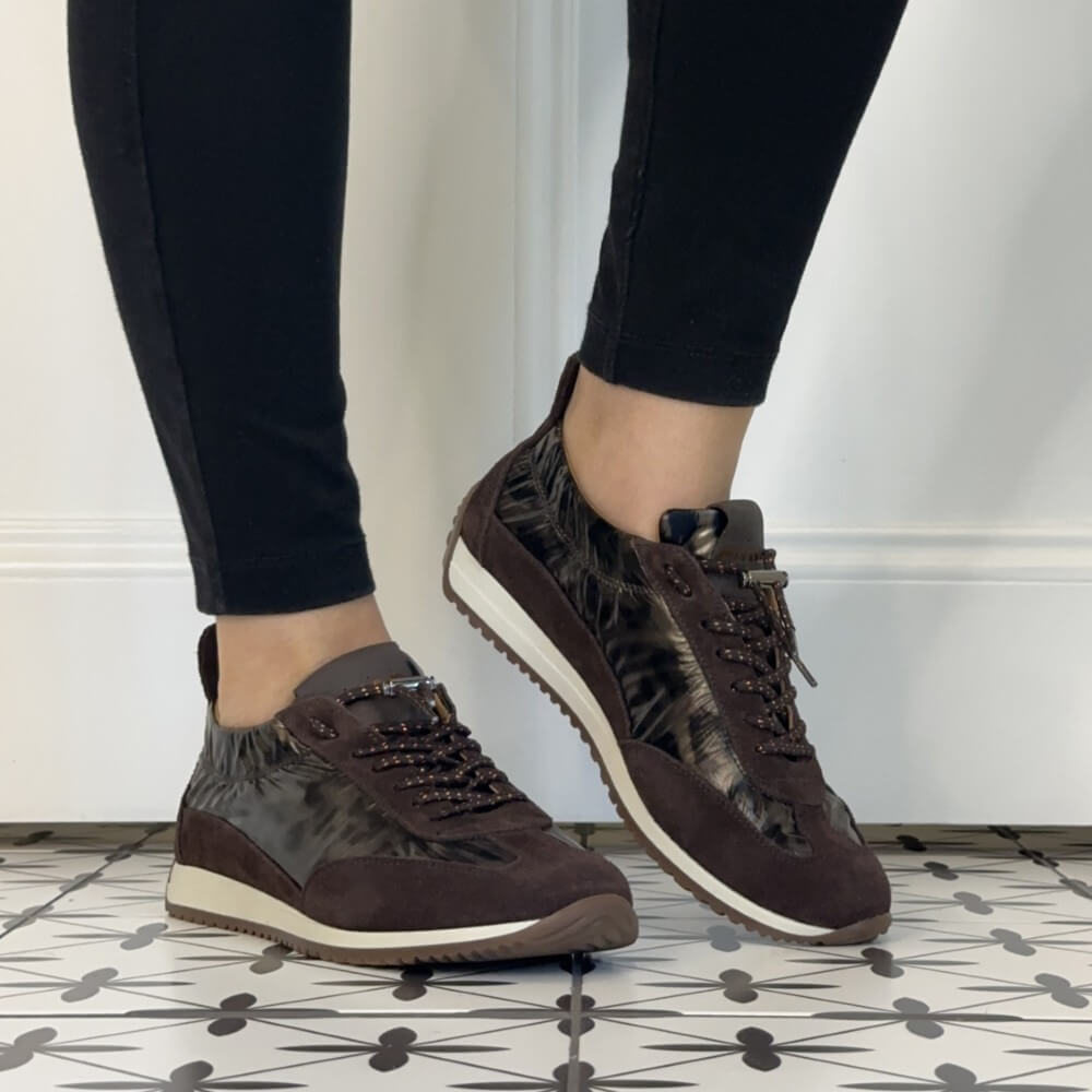A person wearing Meline retro dark brown and beige patterned low-top sneakers with black leggings, standing on a black-and-white patterned tile floor against a white wall.