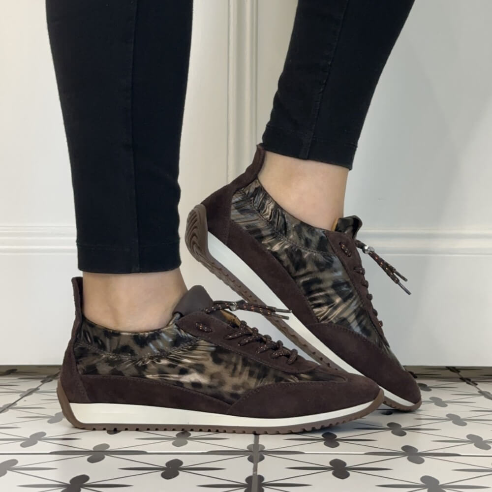 A person wearing Meline retro dark brown and beige patterned low-top sneakers with black leggings, standing on a black-and-white patterned tile floor against a white wall.