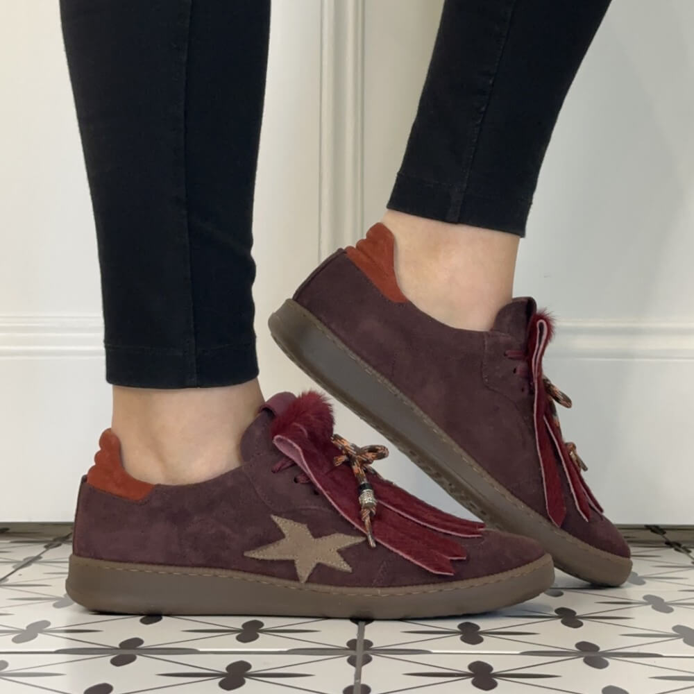 A pair of women's Meline Retro low-top suede sneakers in a deep burgundy colour, shown being worn with black leggings. The shoes feature a beige star on the side, red fringe tassels with beads, and a brown sole.