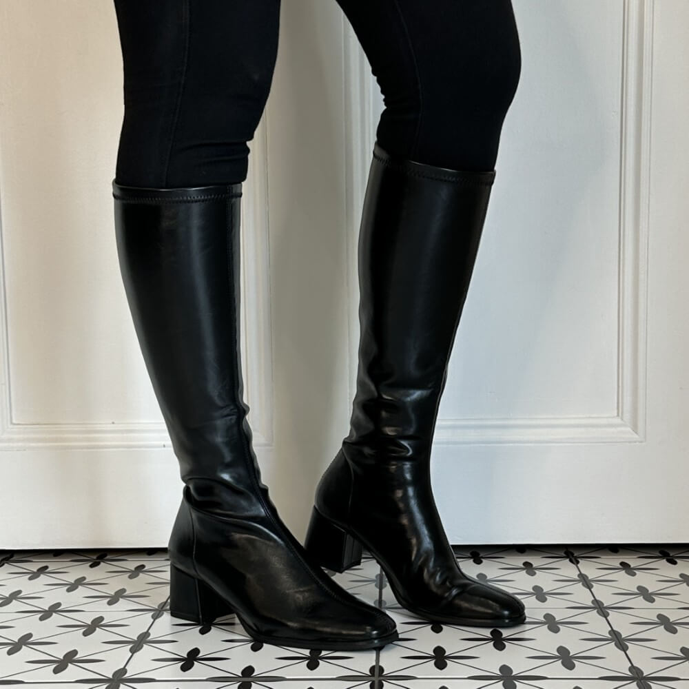 A pair of Miss Elastic black knee-high stretch leather effect boots with block heels, shown on a model wearing black leggings against a white panelled wall and a black-and-white patterned tile floor.