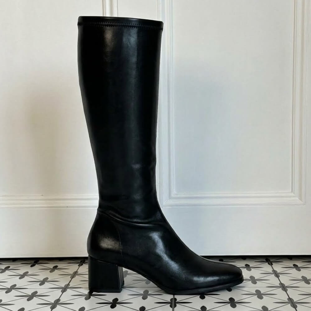 A single black knee-high leather effect boot with a block heel and pointed toe, standing on a black-and-white patterned tile floor in front of a white panelled wall.