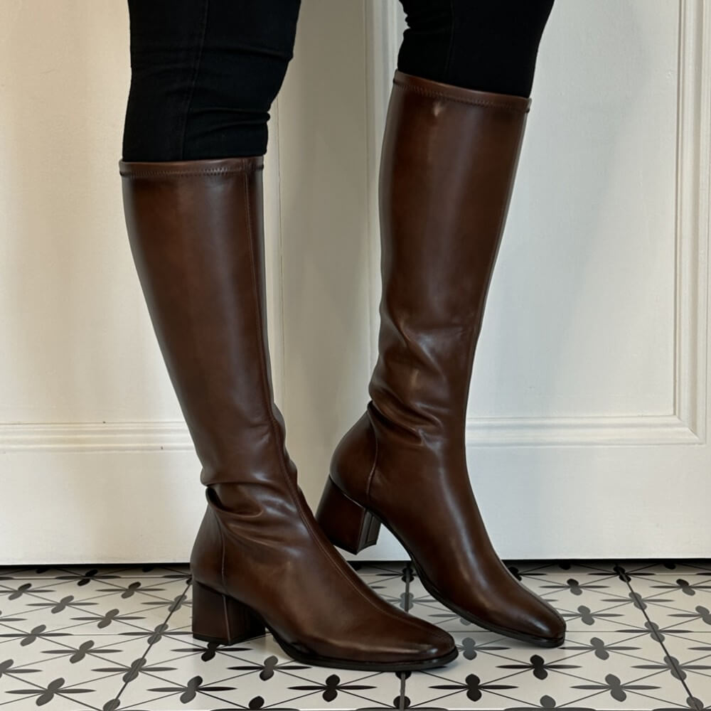 A pair of Miss Elastic dark brown knee-high PU leather boots with block heels, shown on a model wearing black pants against a white panelled door and patterned tile floor.