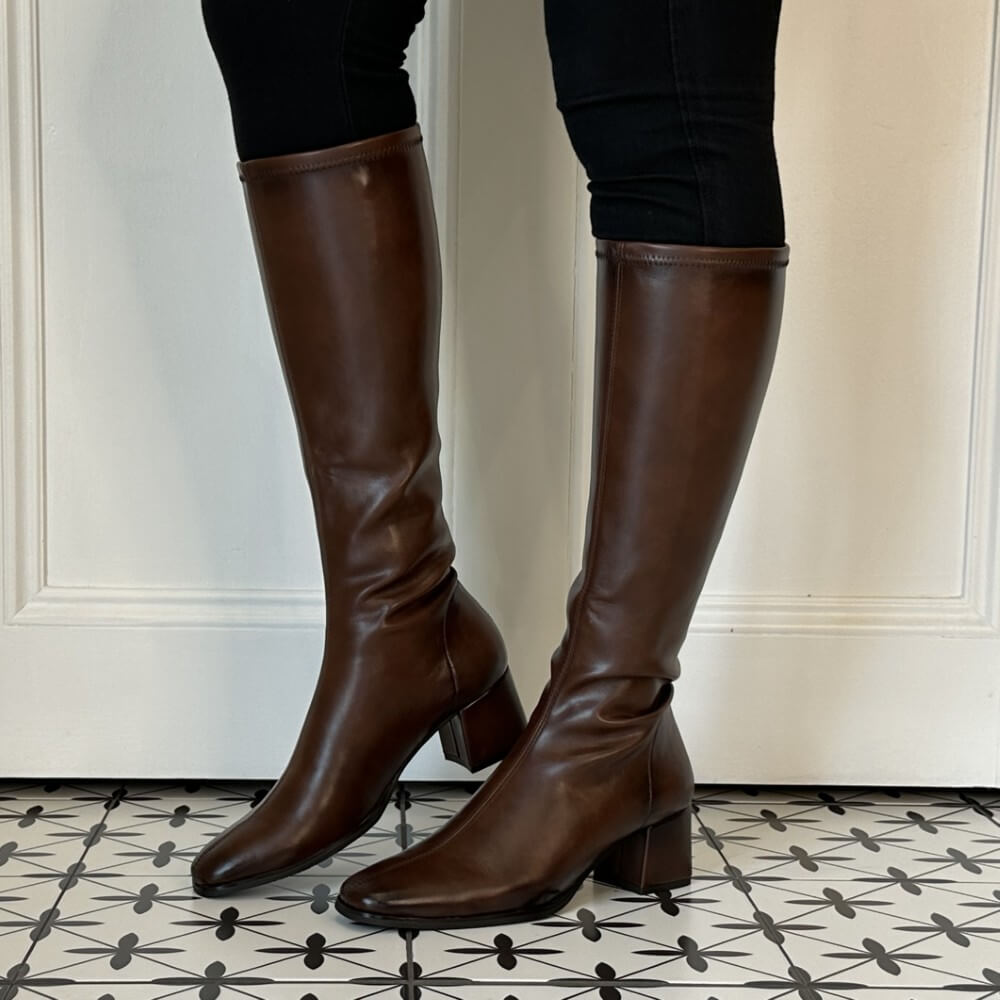 A pair of Miss Elastic dark brown knee-high PU leather boots with block heels, shown on a model wearing black pants against a white panelled door and patterned tile floor.