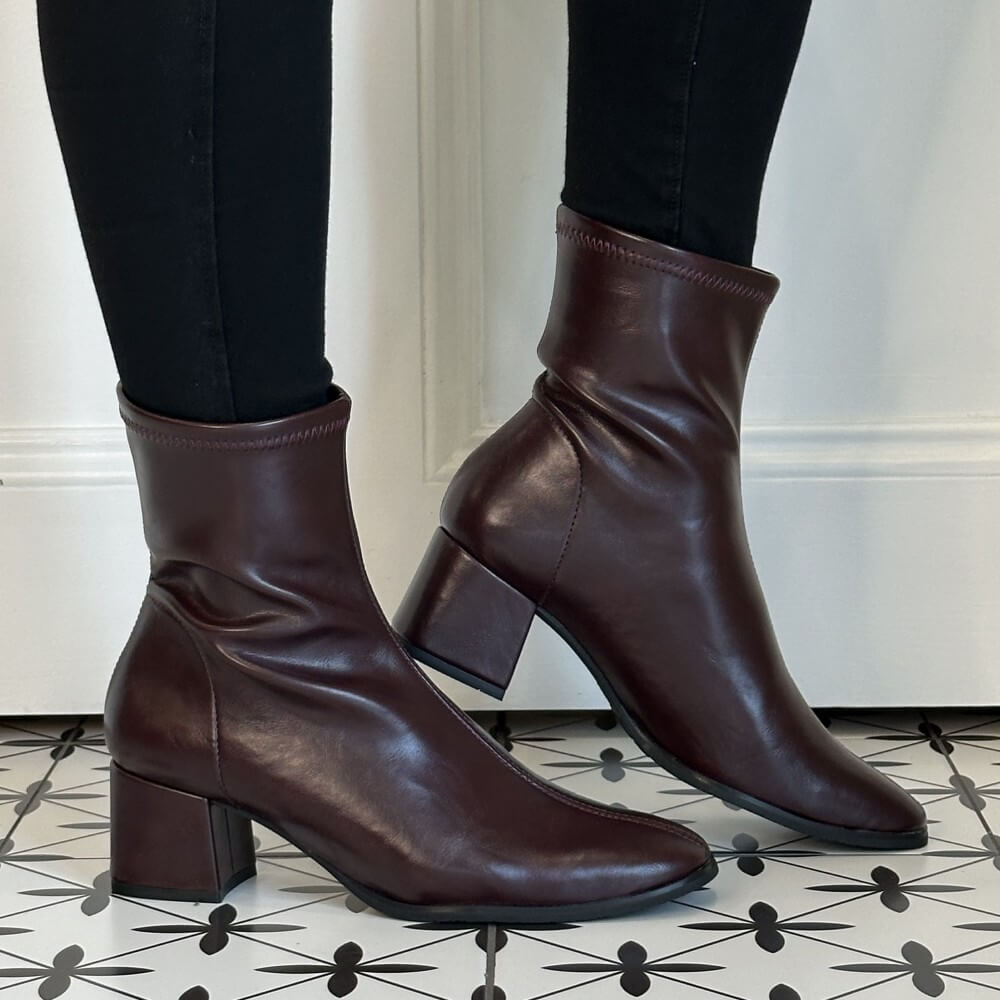 A pair of Miss Elastic Wine ankle boots with block heels, worn with black leggings on a patterned black-and-white tile floor in front of a white wall.