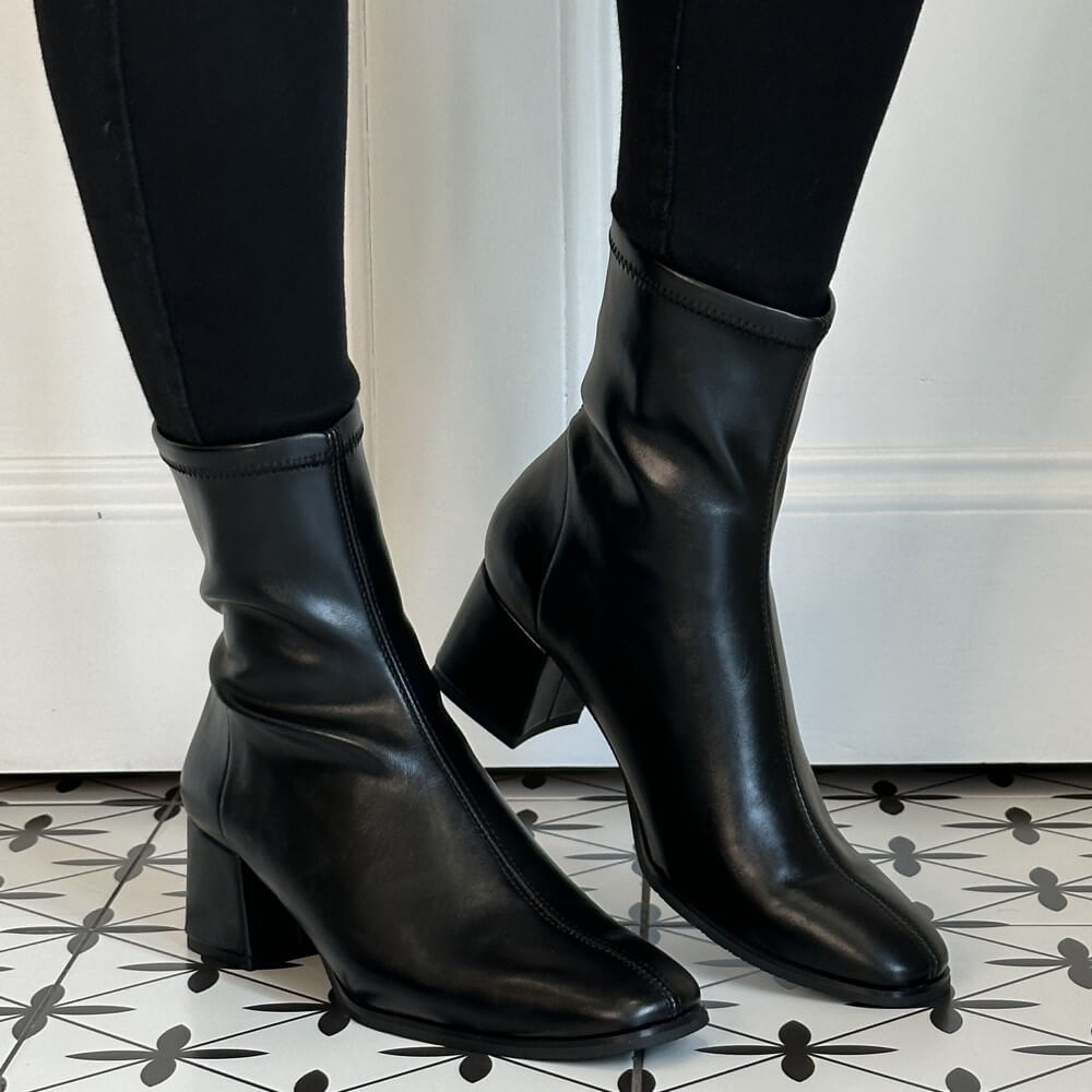A pair of Miss Elastic black ankle boots with block heels, shown on a model wearing black leggings. The boots are positioned on a black and white patterned tile floor against a white wall.