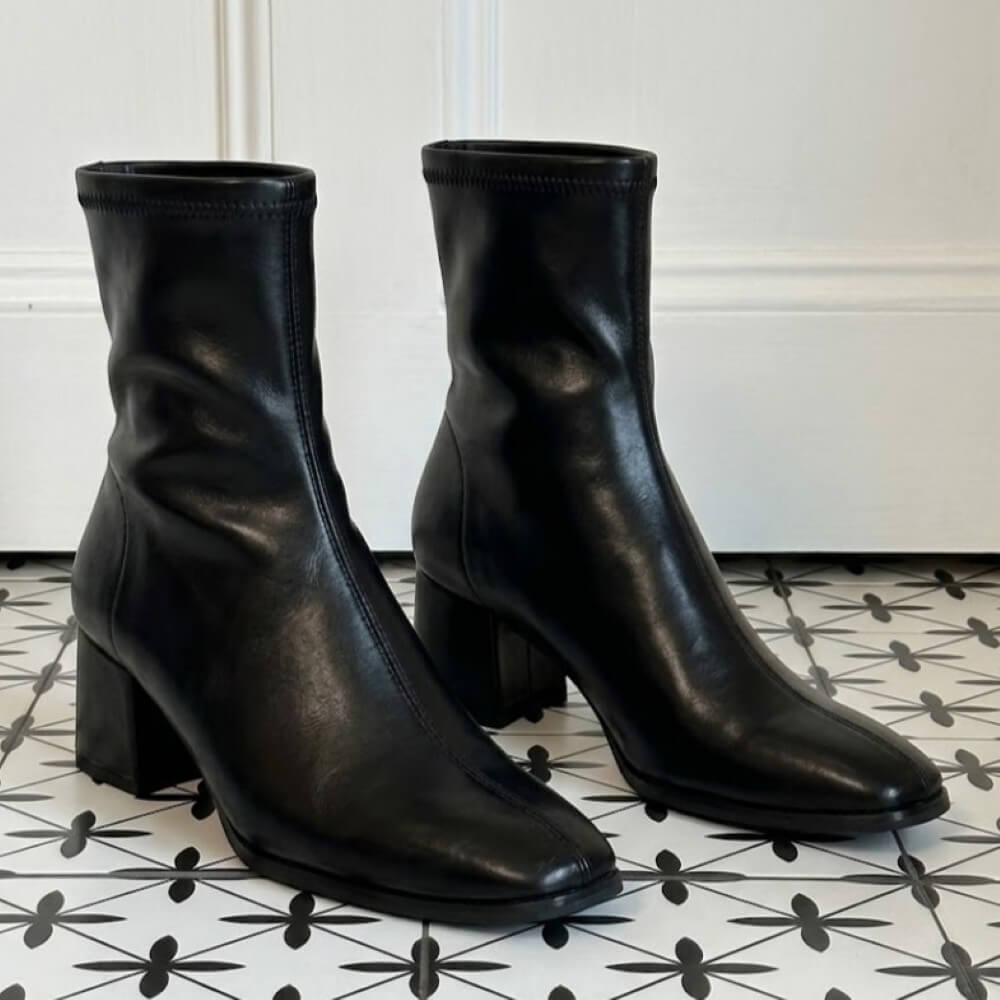  A pair of Miss Elastic black ankle boots with block heels, The boots are positioned on a black and white patterned tile floor against a white wall.