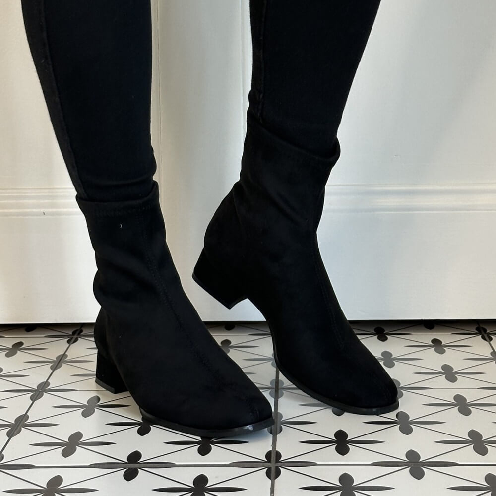 A pair of Miss Elastic black ankle boots with a soft, stretchy fabric upper and low block heels, shown on a model wearing black skinny pants against a white wall and patterned black-and-white floor.