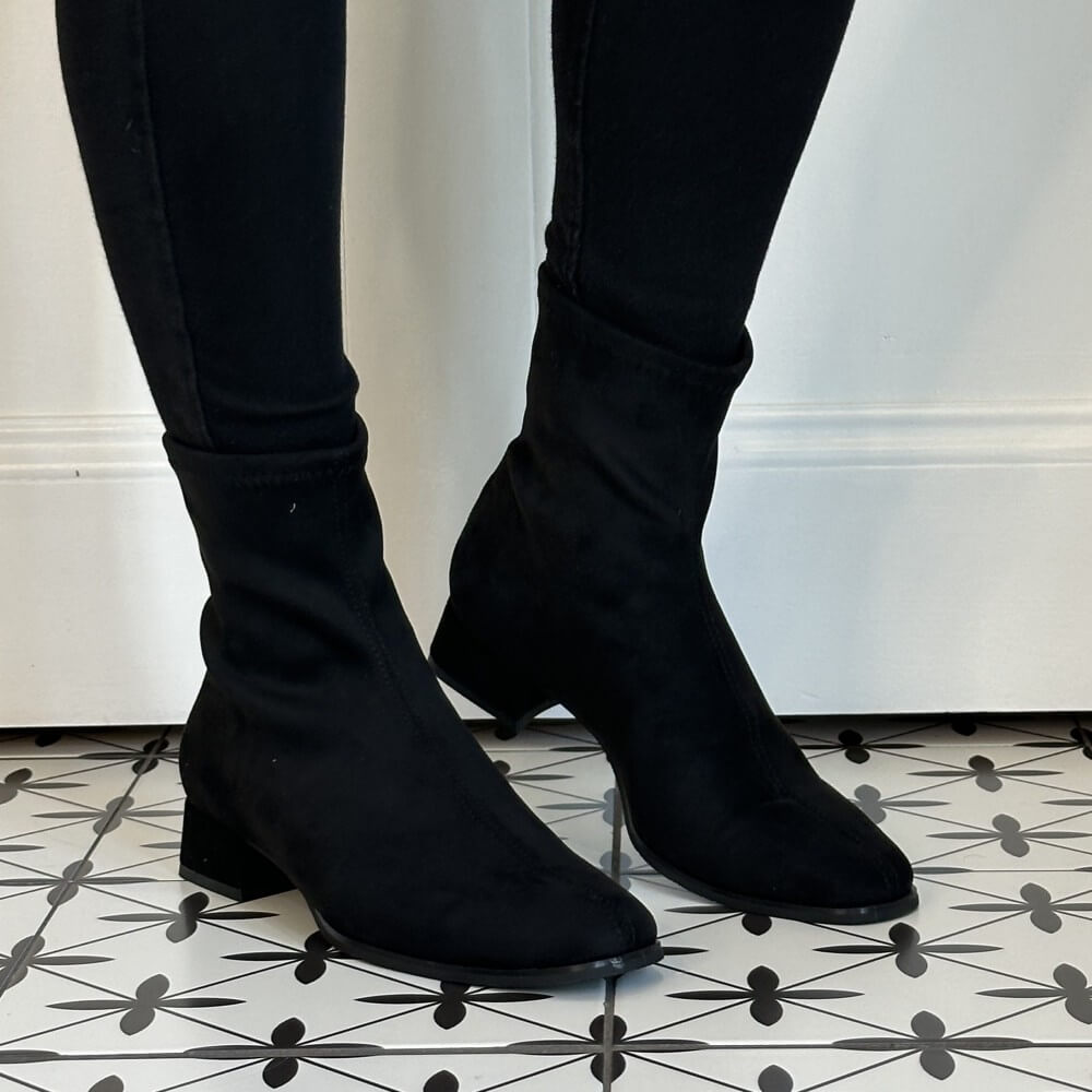 A pair of Miss Elastic black ankle boots with a soft, stretchy fabric upper and low block heels, shown on a model wearing black skinny pants against a white wall and patterned black-and-white floor.