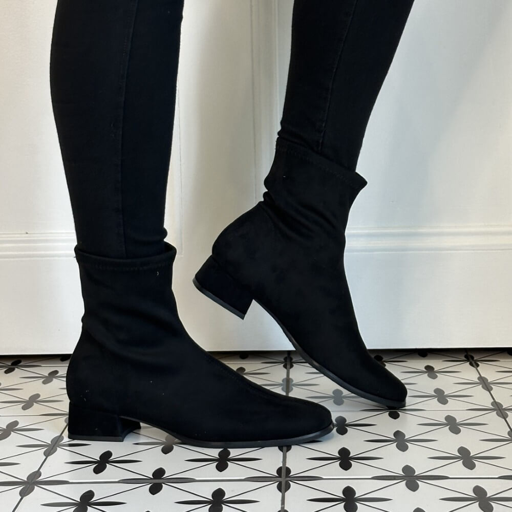 A pair of Miss Elastic black ankle boots with a soft, stretchy fabric upper and low block heels, shown on a model wearing black skinny pants against a white wall and patterned black-and-white floor.