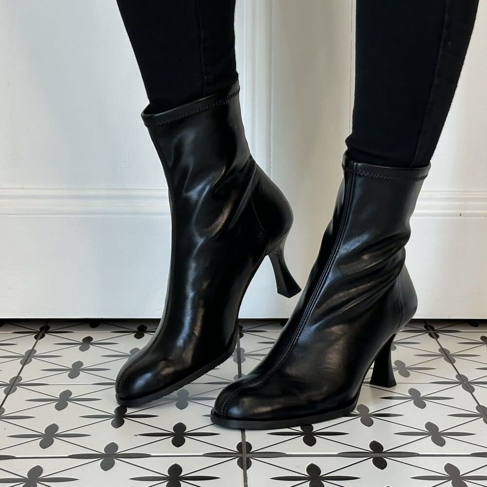 A pair of Miss Elastic black stretch ankle boots with mid-height stiletto heels, worn with black leggings on a black and white patterned tile floor.