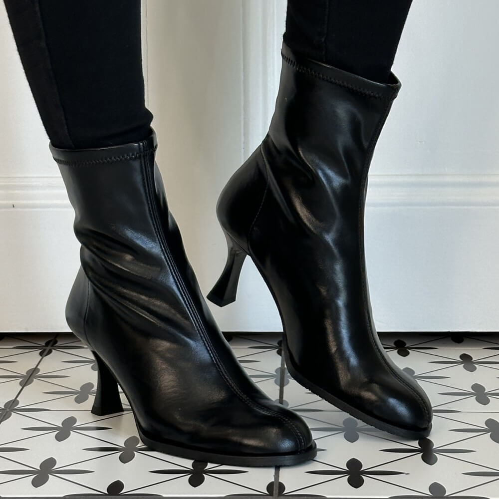 A pair of Miss Elastic black stretch ankle boots with mid-height stiletto heels, worn with black leggings on a black and white patterned tile floor.