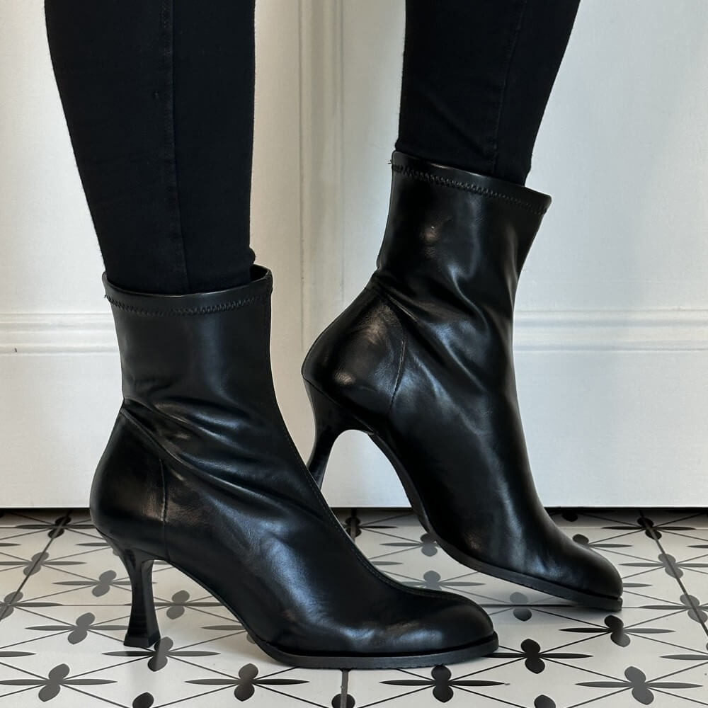 A pair of Miss Elastic black stretch ankle boots with mid-height stiletto heels, worn with black leggings on a black and white patterned tile floor.