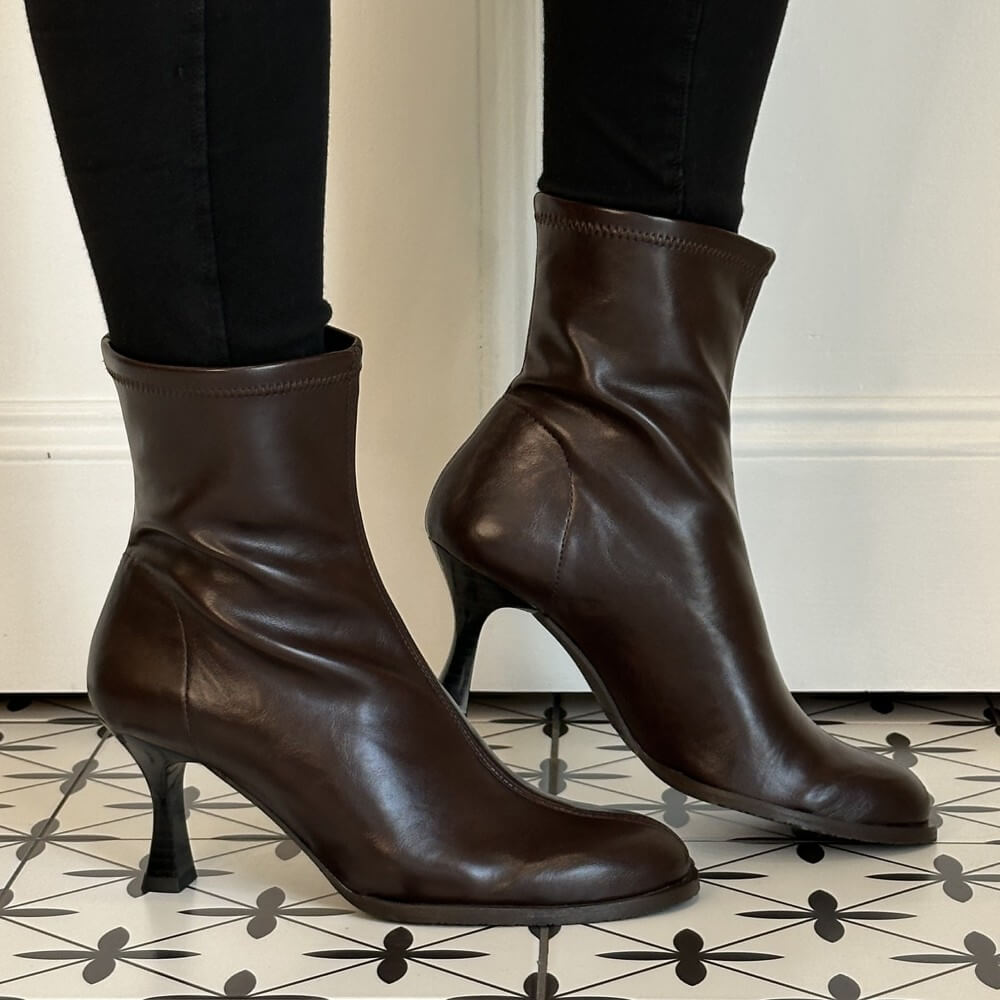 A pair of Miss Elastic dark brown stretch ankle boots with kitten heels, shown on a model wearing black pants. The boots are positioned on a black and white patterned tile floor against a white wall.