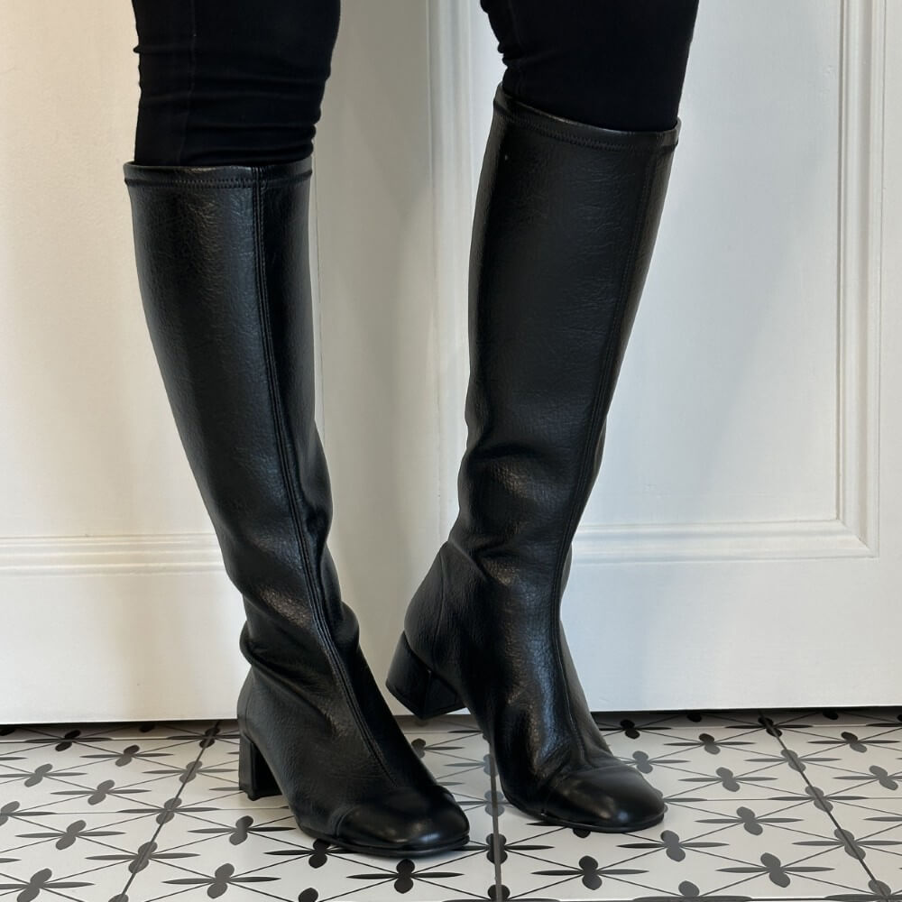 A pair of Miss Elastic black leather knee-high boots with low block heels, shown on a model wearing black leggings against a white panelled door and a black-and-white patterned tile floor.