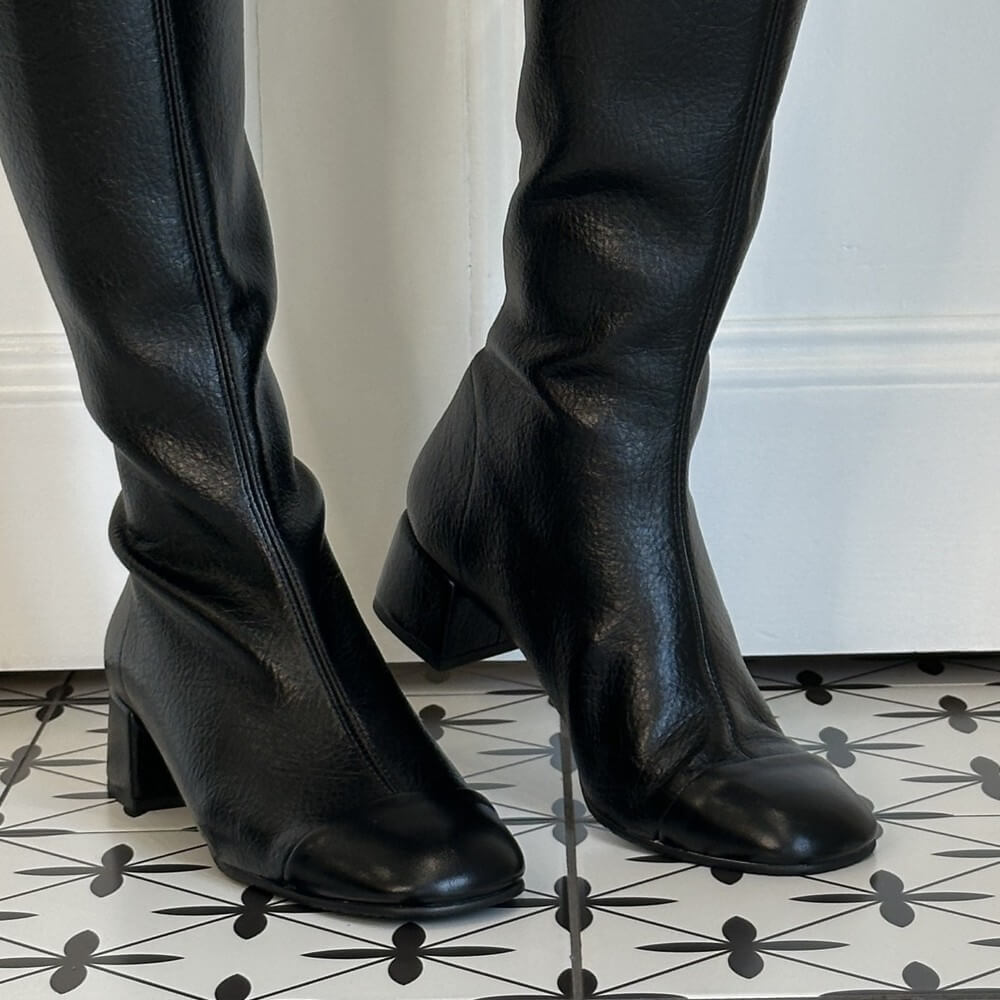 A pair of Miss Elastic black leather knee-high boots with low block heels, shown on a model wearing black leggings against a white panelled door and a black-and-white patterned tile floor.