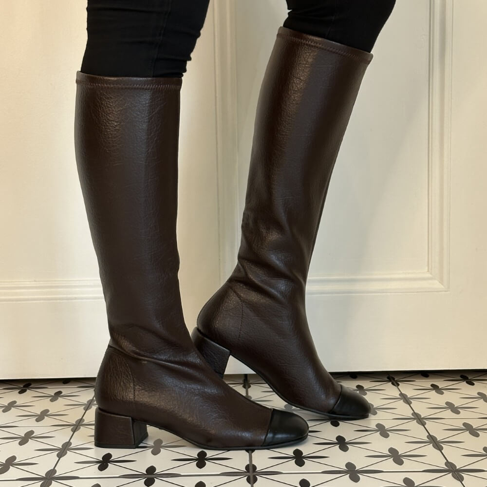A pair of Miss Elastic dark brown knee-high boots with black toe caps, shown on a model wearing black leggings against a white panelled wall and patterned tile floor.