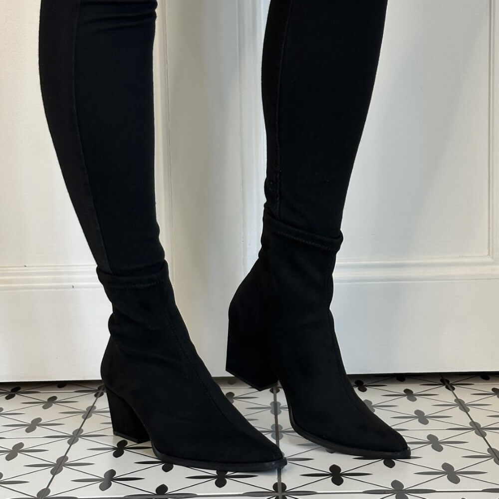 A person wearing a pair of Miss Elastic black stretch ankle boots with block heels, displayed on a black and white patterned tile floor in front of a white panelled wall.