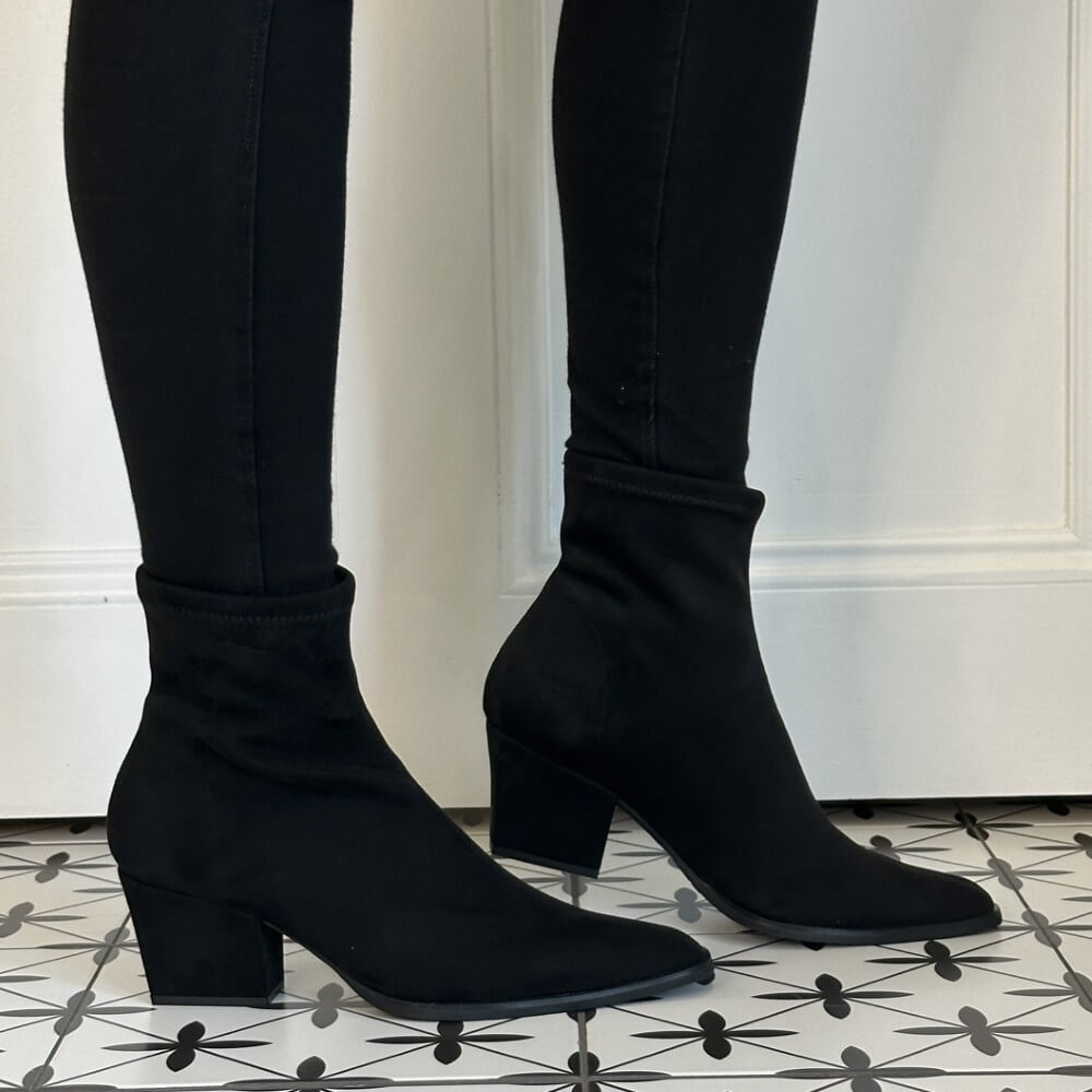 A person wearing a pair of Miss Elastic black stretch ankle boots with block heels, displayed on a black and white patterned tile floor in front of a white panelled wall.