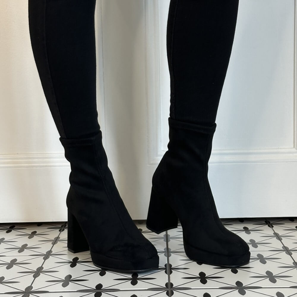 Miss Elastic Black Ankle sock boots with chunky block heels, shown on a model wearing black skinny pants against a white door and patterned tile floor.